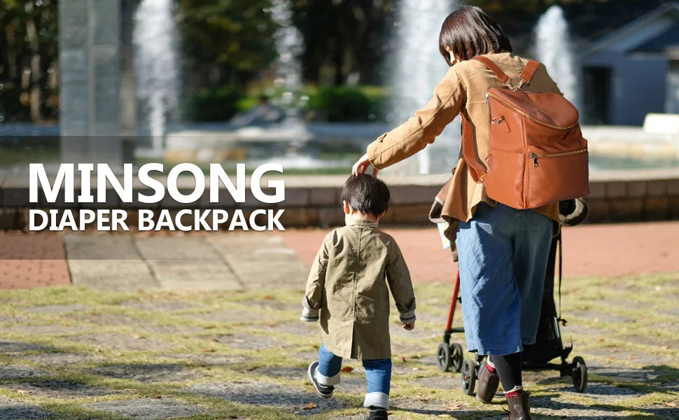 A woman wearing a brown jacket and blue pants carries a Minsong diaper backpack while pushing a stroller in a park.
