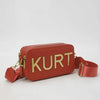 A terracotta red leather pouch bag with gold lettering spelling K U R T and a matching strap.