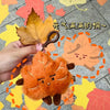 An orange maple leaf plush charm is being held by a hand against a background of colorful autumn leaves.