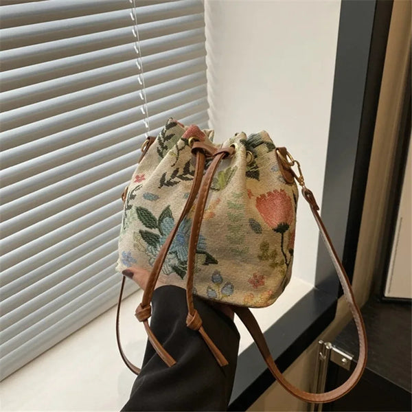 A beige floral drawstring bucket bag with brown leather straps is shown against a white window blind.