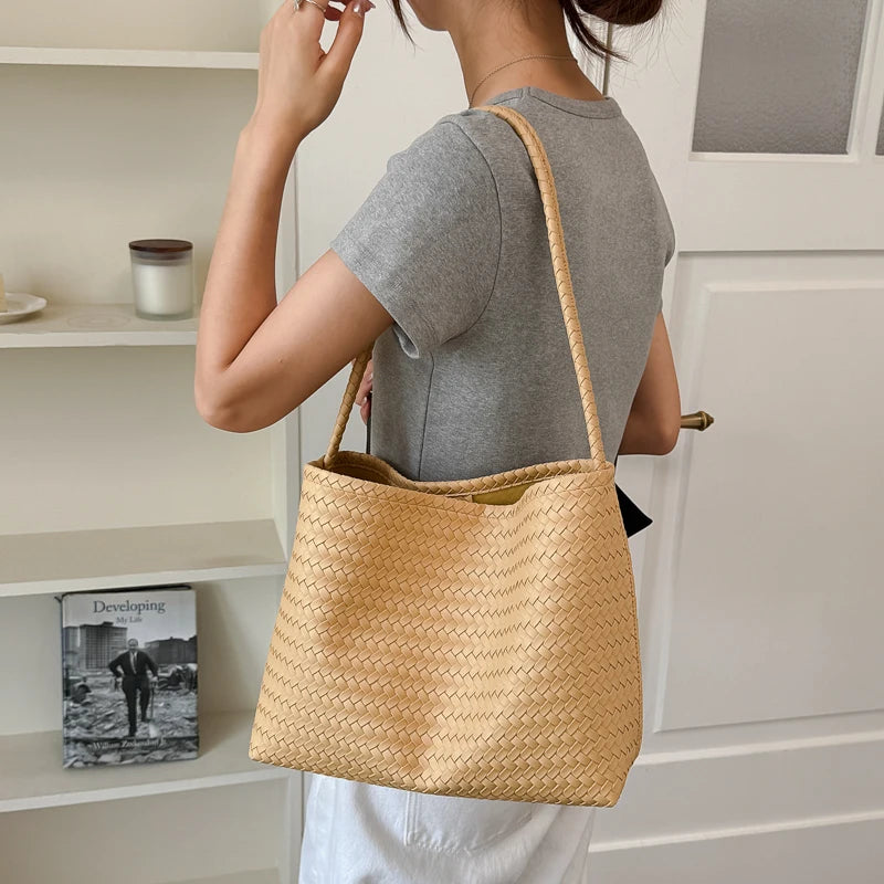 A woman holds a tan woven bucket shoulder bag against a white cabinet with shelves.