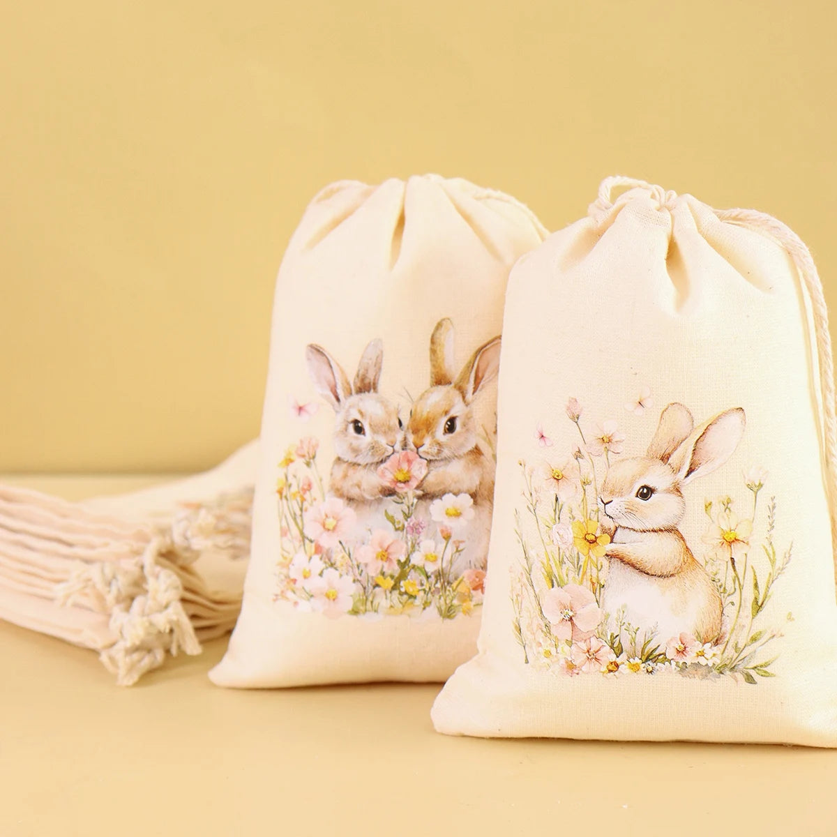 A set of six light beige drawstring gift bags featuring watercolor illustrations of baby rabbits and flowers.