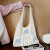 A white knitted tote bag with light blue embroidery is being held by a person against a wooden background.