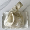 A beige embroidered silk evening clutch with a chain strap resting on a white decorative plate.