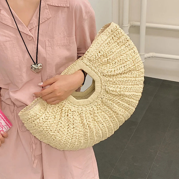 Eine Person hält eine cremefarbene Rattan-Halbmond-Strandtasche vor einem rosa Hemd.