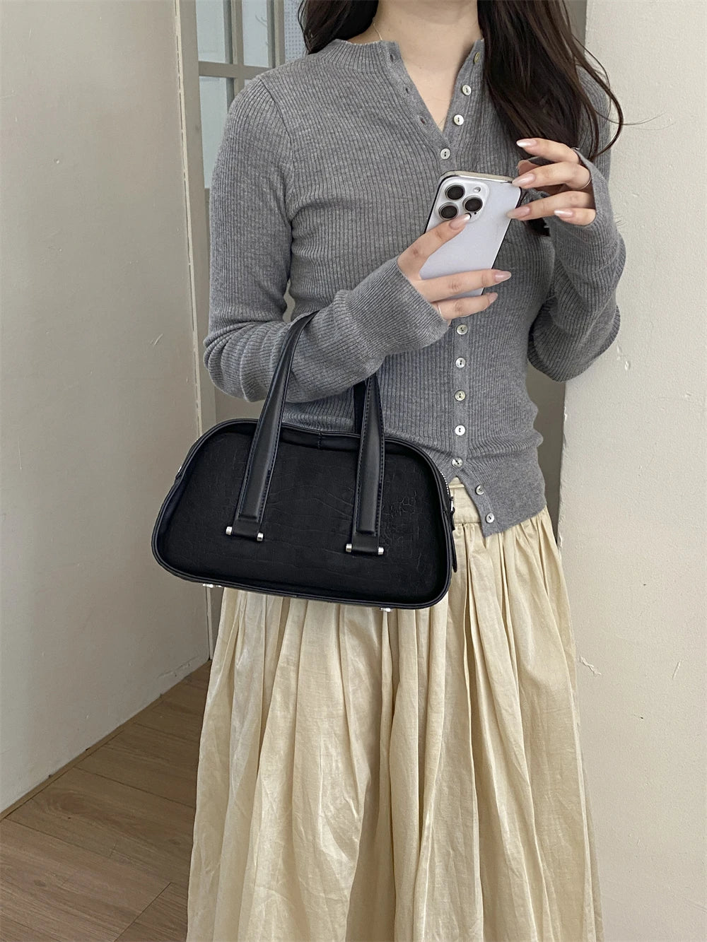 A woman wearing a gray sweater and light skirt holds a black crocodile textured mini crossbody bag and a smartphone.