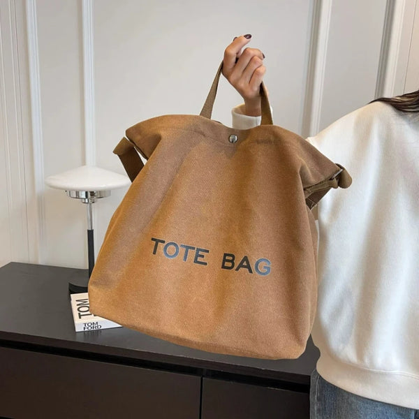 A person holds a brown canvas tote bag with the text TOTE BAG on it against a white background.
