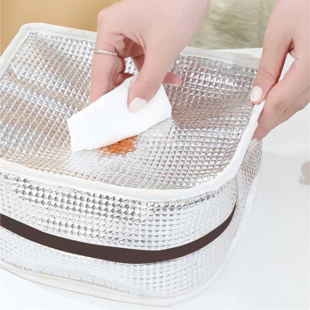 A hand is cleaning a square, silver-textured insulated lunch bag with a white cloth inside.
