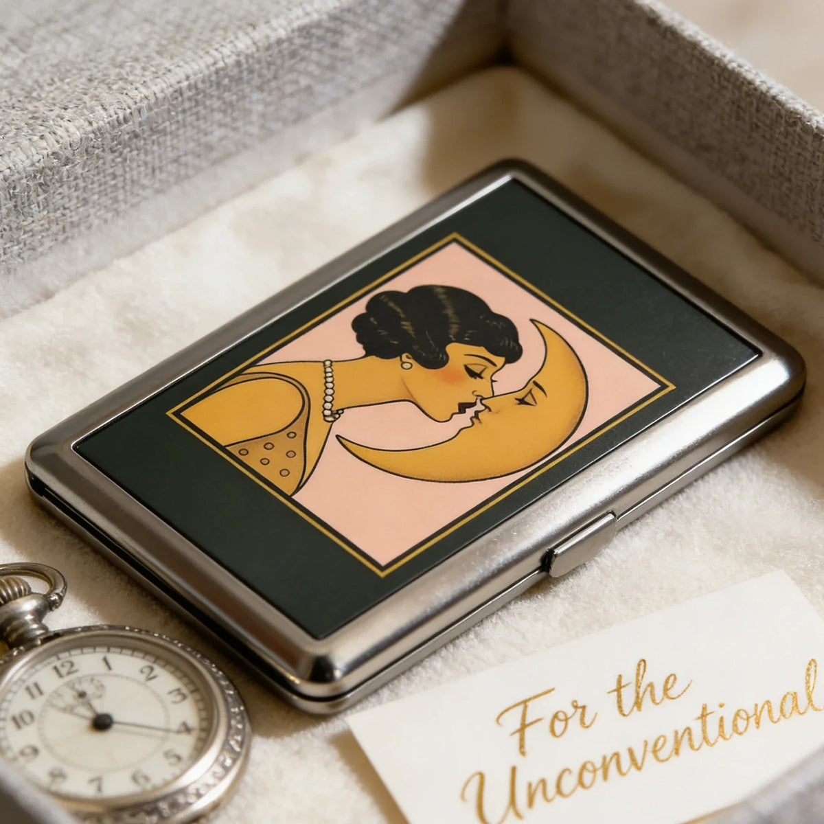 A silver metal card holder featuring an Art Deco illustration of a woman kissing the moon on a pink background.