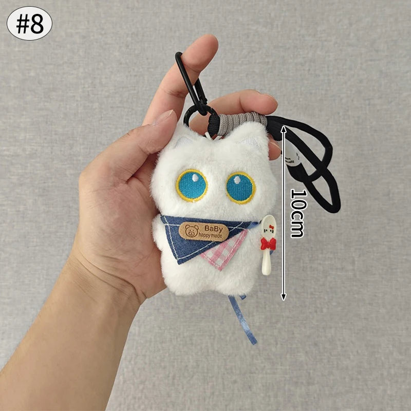 Colgante de peluche de gato blanco con ojos azules y un lazo azul, sostenido por una mano.