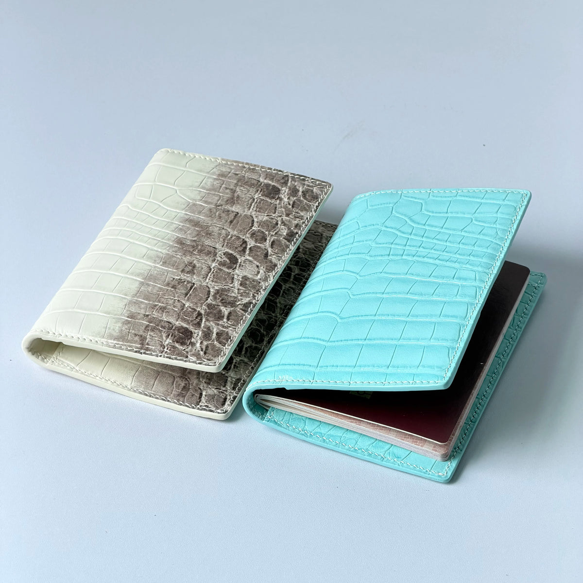 A stack of genuine leather passport holders featuring crocodile patterns in beige, gray, and turquoise colors.