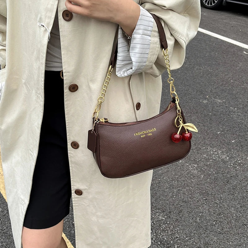 A brown leather shoulder bag with a gold chain and cherry pendant is carried by a person wearing a beige trench coat.