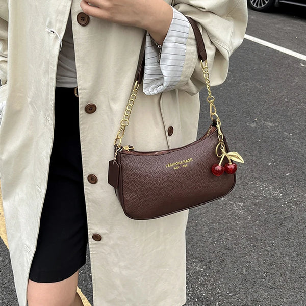 A brown leather shoulder bag with a gold chain and cherry pendant is carried by a person wearing a beige trench coat.