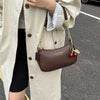 A brown leather shoulder bag with a gold chain and cherry pendant is carried by a person wearing a beige trench coat.