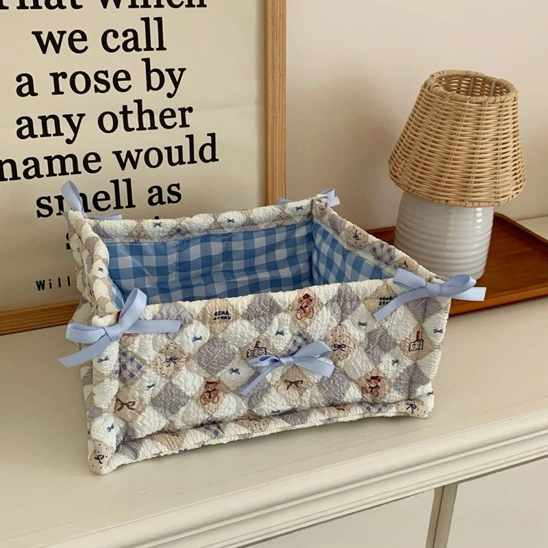 A quilted cotton storage basket with a lace flower design and light blue ribbon, sitting on a wooden surface.