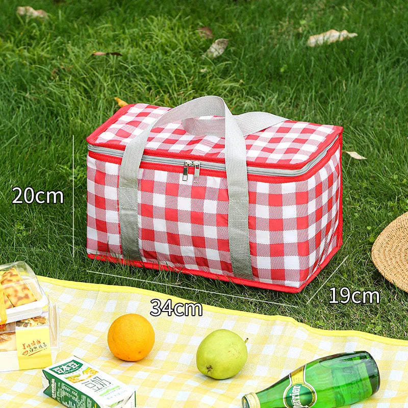 A red and white checkered insulated picnic tote bag lying on green grass with fruit and a bottle.