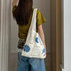 A person wearing a olive green shirt and blue jeans holds a white knitted tote bag with blue floral and star embroidery.