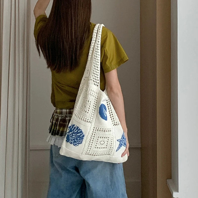 A person wearing a olive green shirt and blue jeans holds a white knitted tote bag with blue floral and star embroidery.