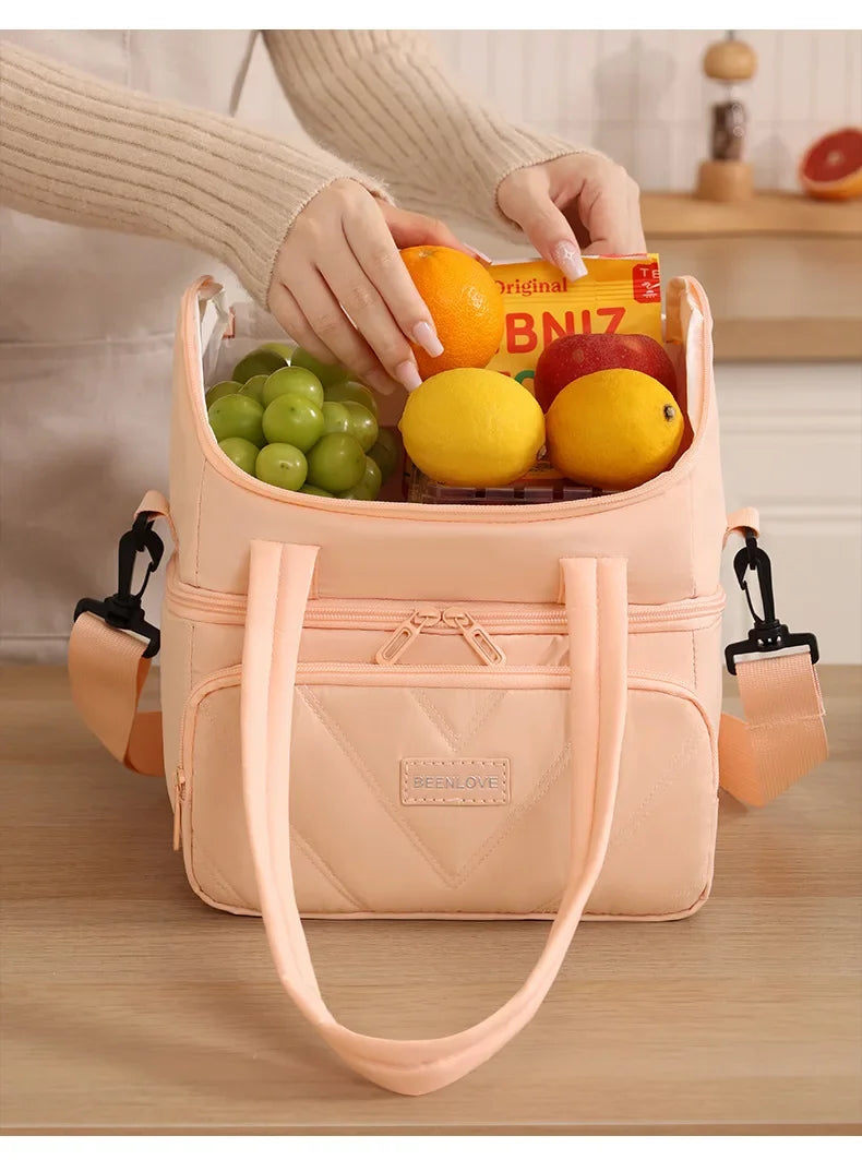 A light pink insulated lunch bag is shown being held by a hand, filled with fresh fruit like oranges and apples.