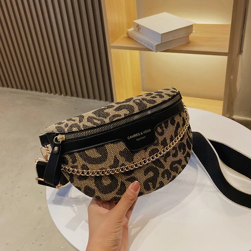 Une main tient un sac bandoulière à taille imprimé léopard noir et or sur une surface blanche.