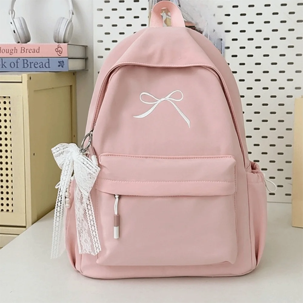 A pink embroidered backpack with a white bow and pocket on a white surface.