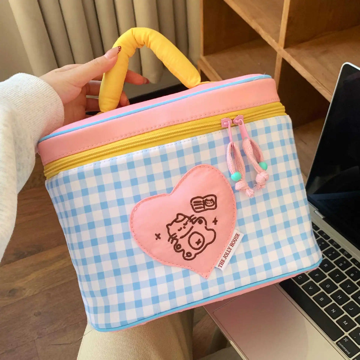 A pink and blue checkered insulated lunch bag with a heart design and a yellow handle is held by a hand.
