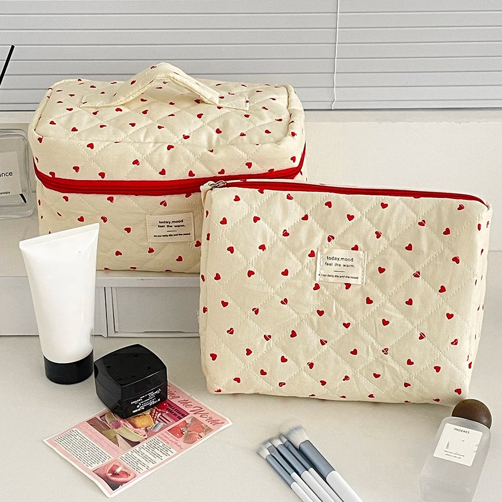 A cream quilted makeup bag with a red trim and a pattern of small red hearts, displayed next to a white tube.