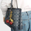 A dark blue leather handbag with gold hardware and a crystal fruit charm keychain is carried by a person wearing a white shir