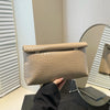 Un bolso de mano tejido beige se muestra sobre una superficie de vidrio junto a un espejo de marco plateado.
