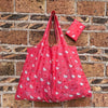 A pink reusable shopping bag with a white cat pattern is displayed against a brick wall.
