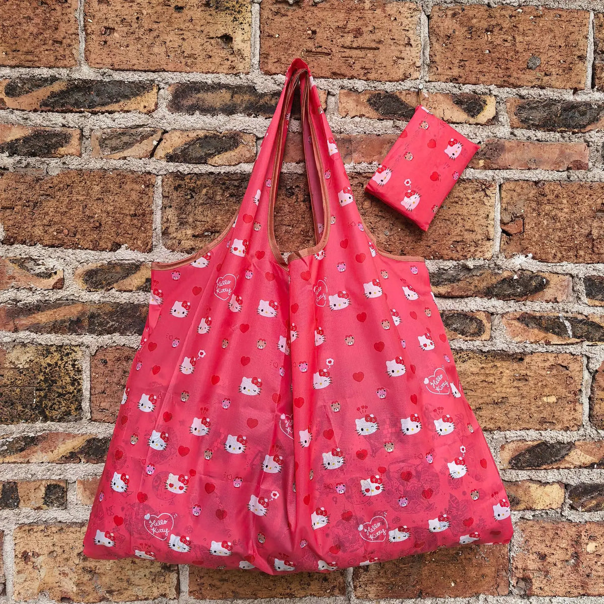 A pink reusable shopping bag with a white cat pattern is displayed against a brick wall.