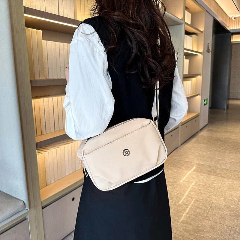 Una mujer con vestido negro y camisa blanca lleva un bolso cruz de nylon impermeable beige en un pasillo bien iluminado.