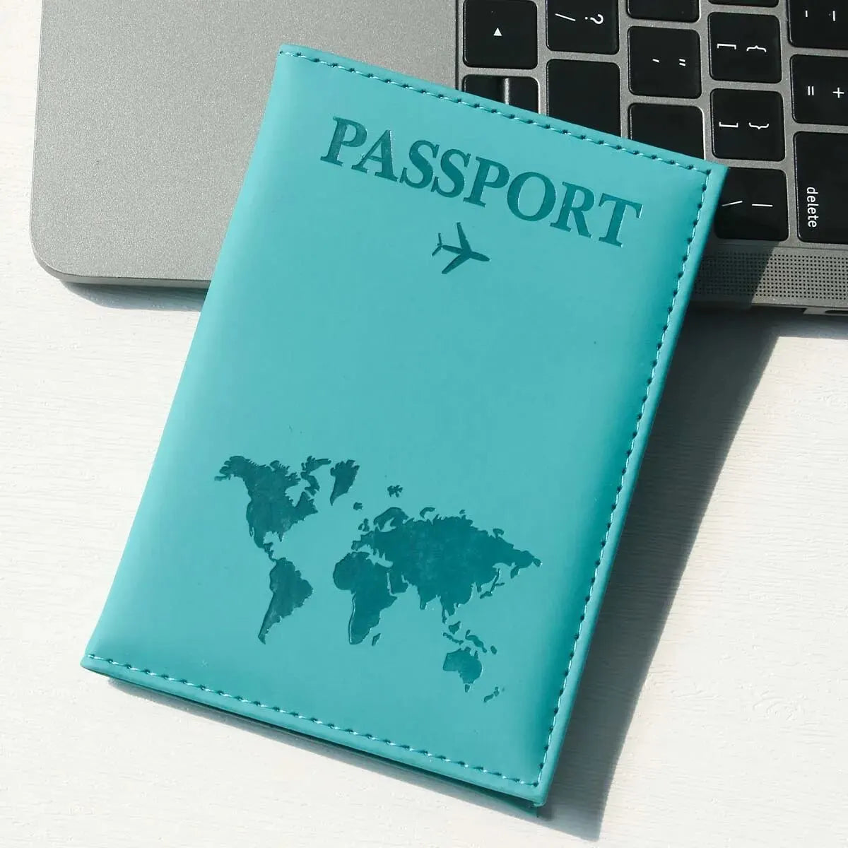 A teal leather passport holder with a world map design resting on a white surface next to a laptop.