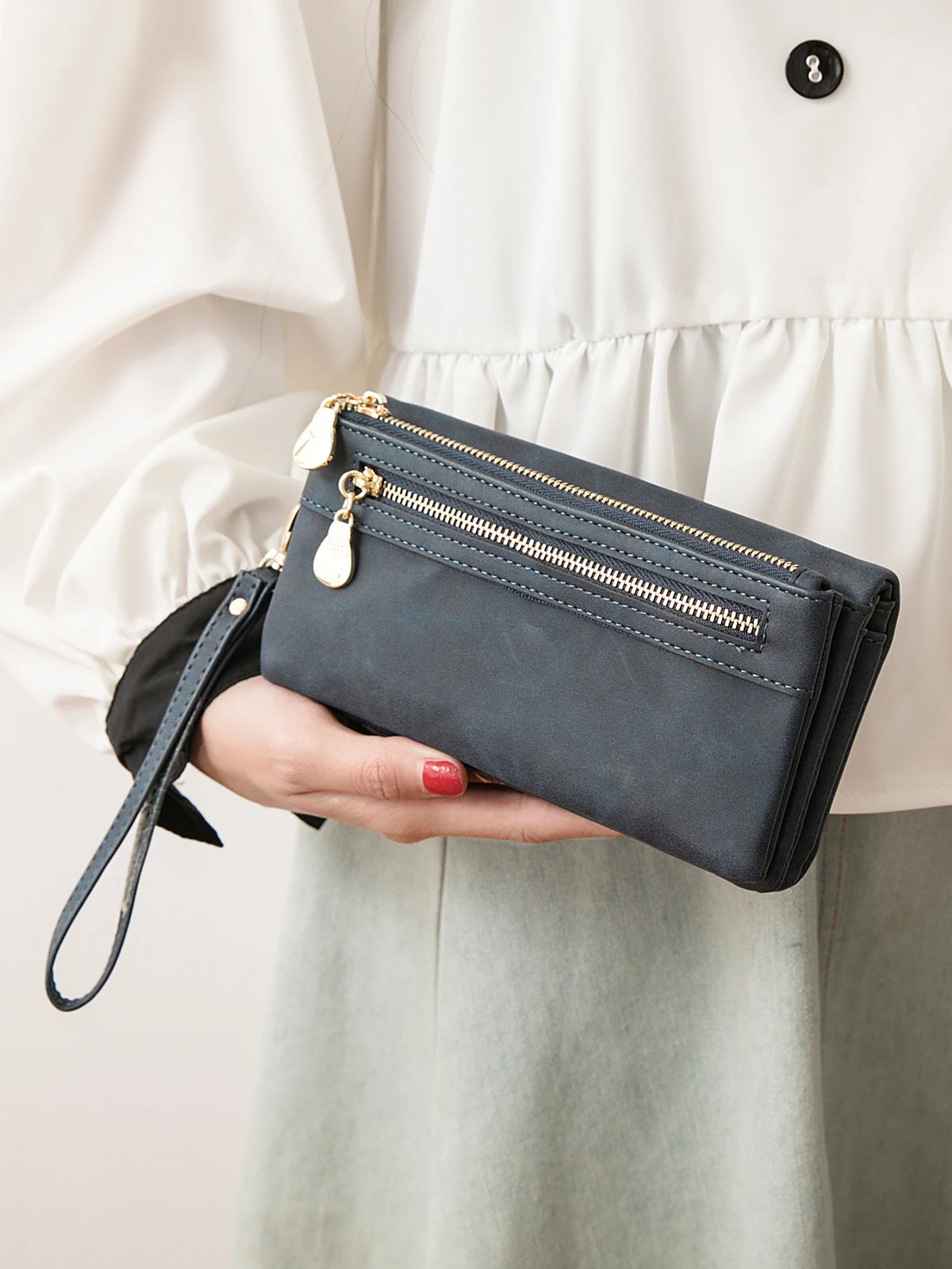 A hand holds a dark gray leather wallet with a gold zipper and a leather tag against a light colored blouse.
