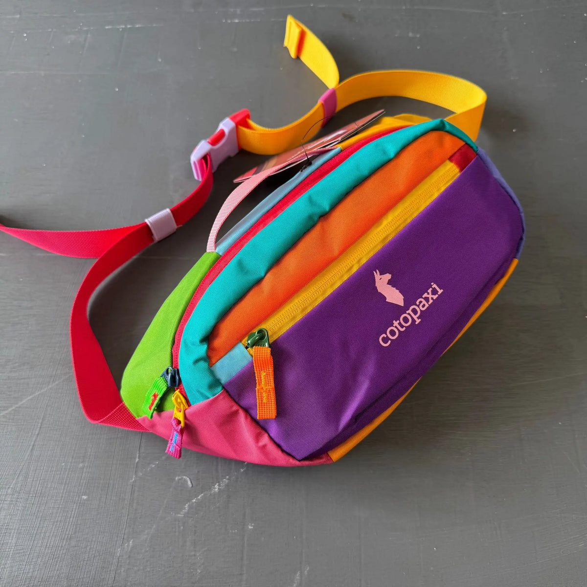 Bunte, straphaltige Outdoor-Crossbody-Tasche mit farbigen Stoffpaneelen und Trageriem auf grauem Untergrund.