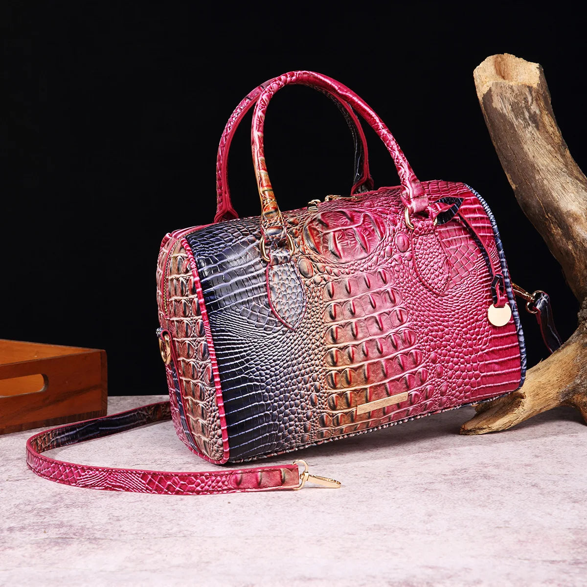 A red crocodile pattern luxury leather crossbody tote bag resting on a light surface next to a piece of driftwood.