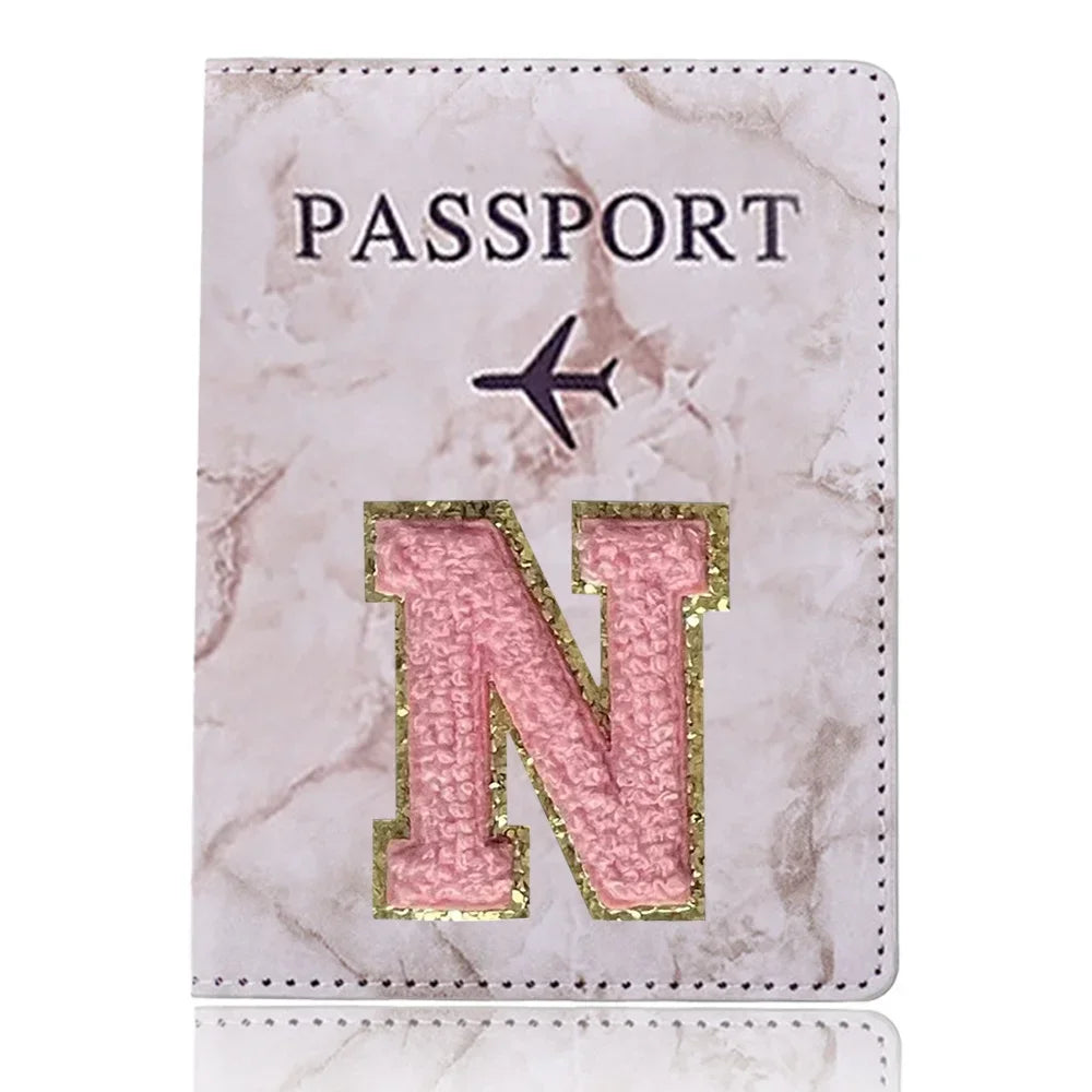 Portafolio de pasaporte de mármol rosa con una inicial N dorada y un símbolo de avión negro.