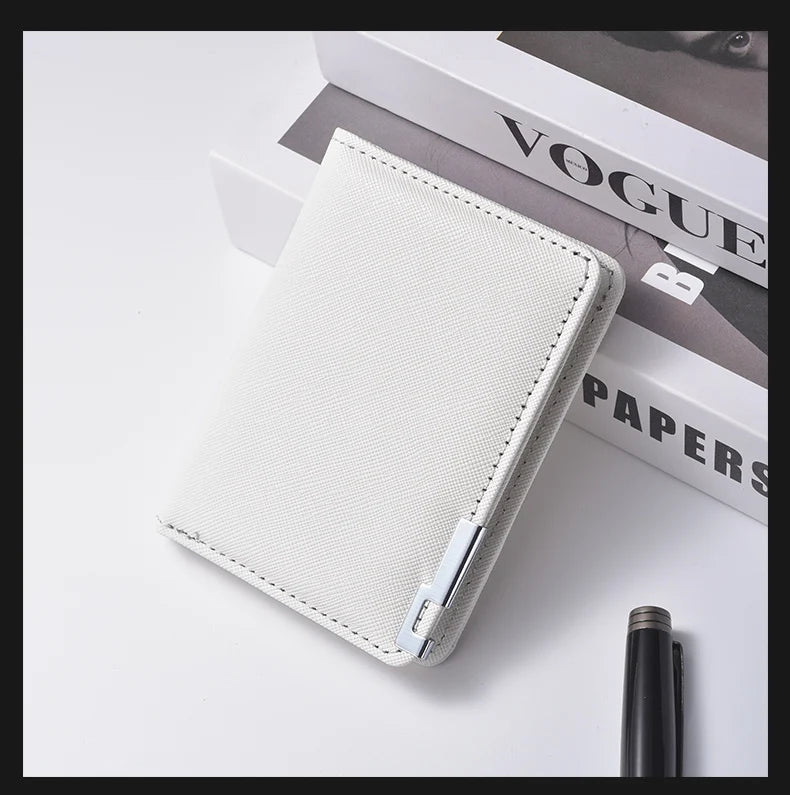 A white slim vertical men's card wallet resting on a white surface next to a black pen and a Vogue magazine.