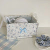 A white quilted cotton storage basket with a lace flower design holds a white ball of yarn.