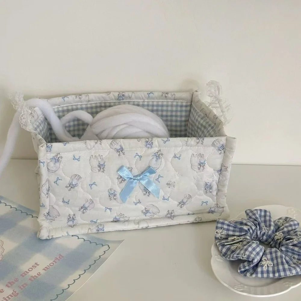 A white quilted cotton storage basket with a lace flower design holds a white ball of yarn.