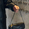 A hand wearing a black leather jacket holds a black vintage cow leather cylinder shoulder bag against a textured gray wall.