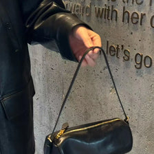 A hand wearing a black leather jacket holds a black vintage cow leather cylinder shoulder bag against a textured gray wall.