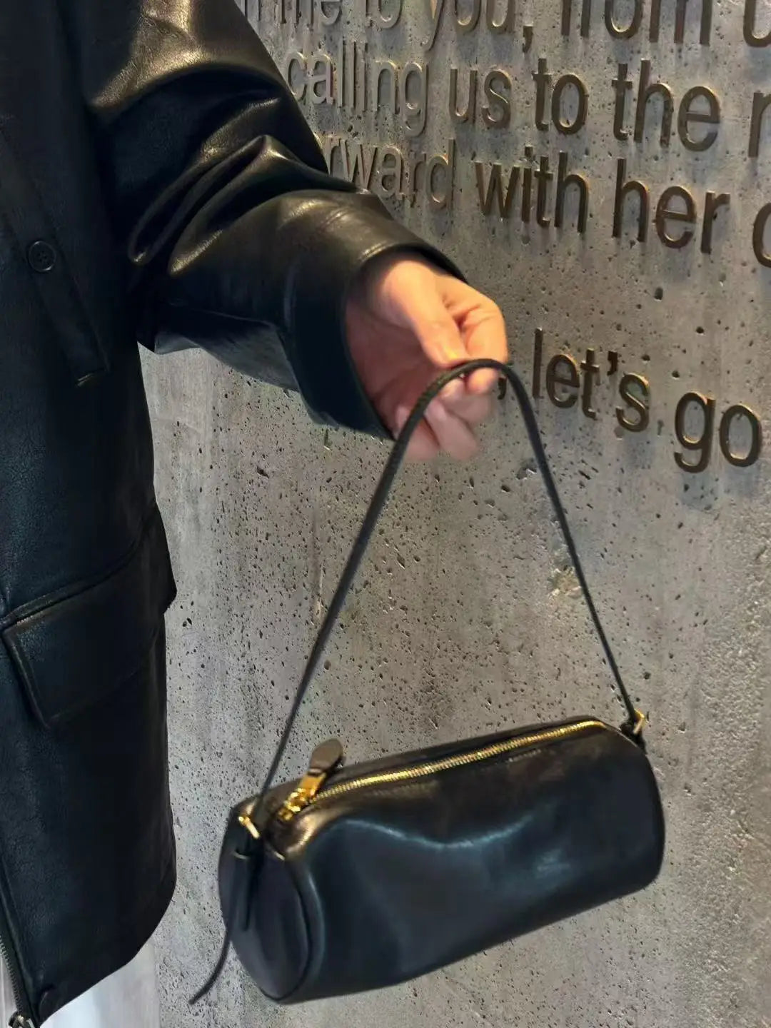 A hand wearing a black leather jacket holds a black vintage cow leather cylinder shoulder bag against a textured gray wall.
