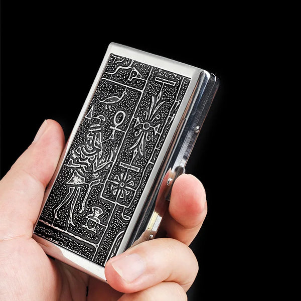 Une main tient un porte-cigarette en métal argenté gravé de symboles anciens sur fond noir.