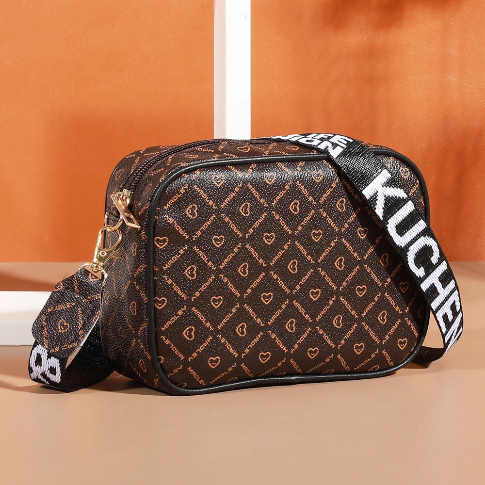 A brown patterned fashion crossbody shoulder bag with black trim and white lettering is displayed on a light surface.