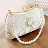A light beige clutch bag with white pearl detailing and gold hardware resting on a wooden surface.