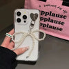 A hand holds a clear phone case featuring a pearl bowknot keychain with a heart charm against a pink background.