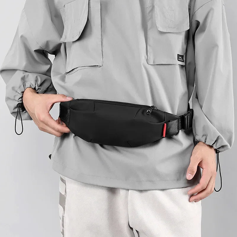 A person holds a black water-resistant nylon waist bag with an adjustable strap against a gray jacket.