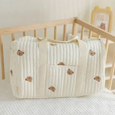 A cream-colored quilted diaper bag tote with a flap and pocket is shown resting on a white surface.