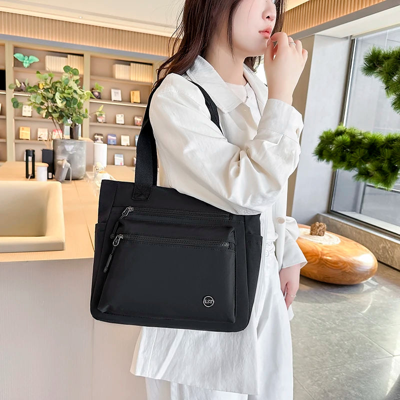 A woman holds a black large capacity waterproof canvas tote bag in a light-colored outfit indoors.